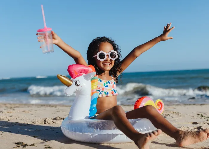Cheerful young girl with sunglasses and unicorn float relaxing at the beach, holding a drink, and having fun during a summer vacation.